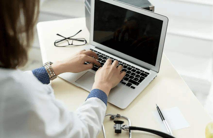 Lekarka siedzi przy biurku i pisze na klawiaturze, z prawej strony na biurku znajduje się stetoskop i długopis, z prawej okulary.