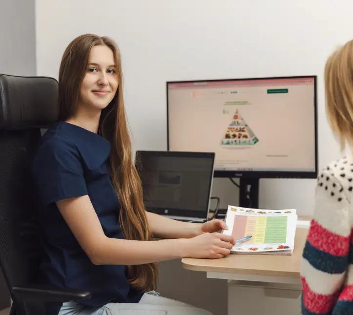 Uśmiechnięta kobieta z długimi włosami, która przeprowadza konsultacje dietetyczne z pacjentką, pokazuje plan żywienia.