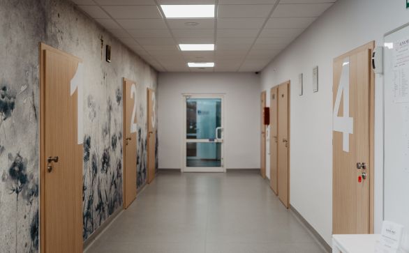 Korytarz przychodni Alma Med z drzwiami w kolorze drewna, ściana z lewej strony pokryta jest tapetą z roślinnym motywem w monochromatycznych kolorach, reszta ścian w kolorze białym