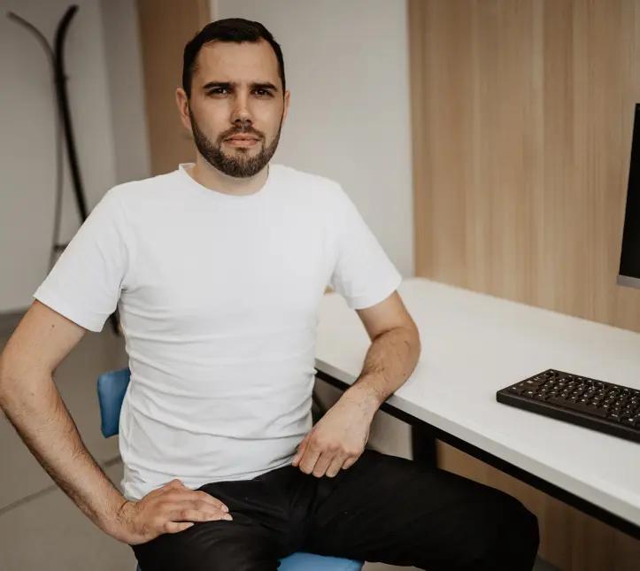 Ciemnowłosy mężczyzna z brodą w białej koszulce i czarnych spodniach, siedzący na krześle przy komputerze w gabinecie fizjoterapii